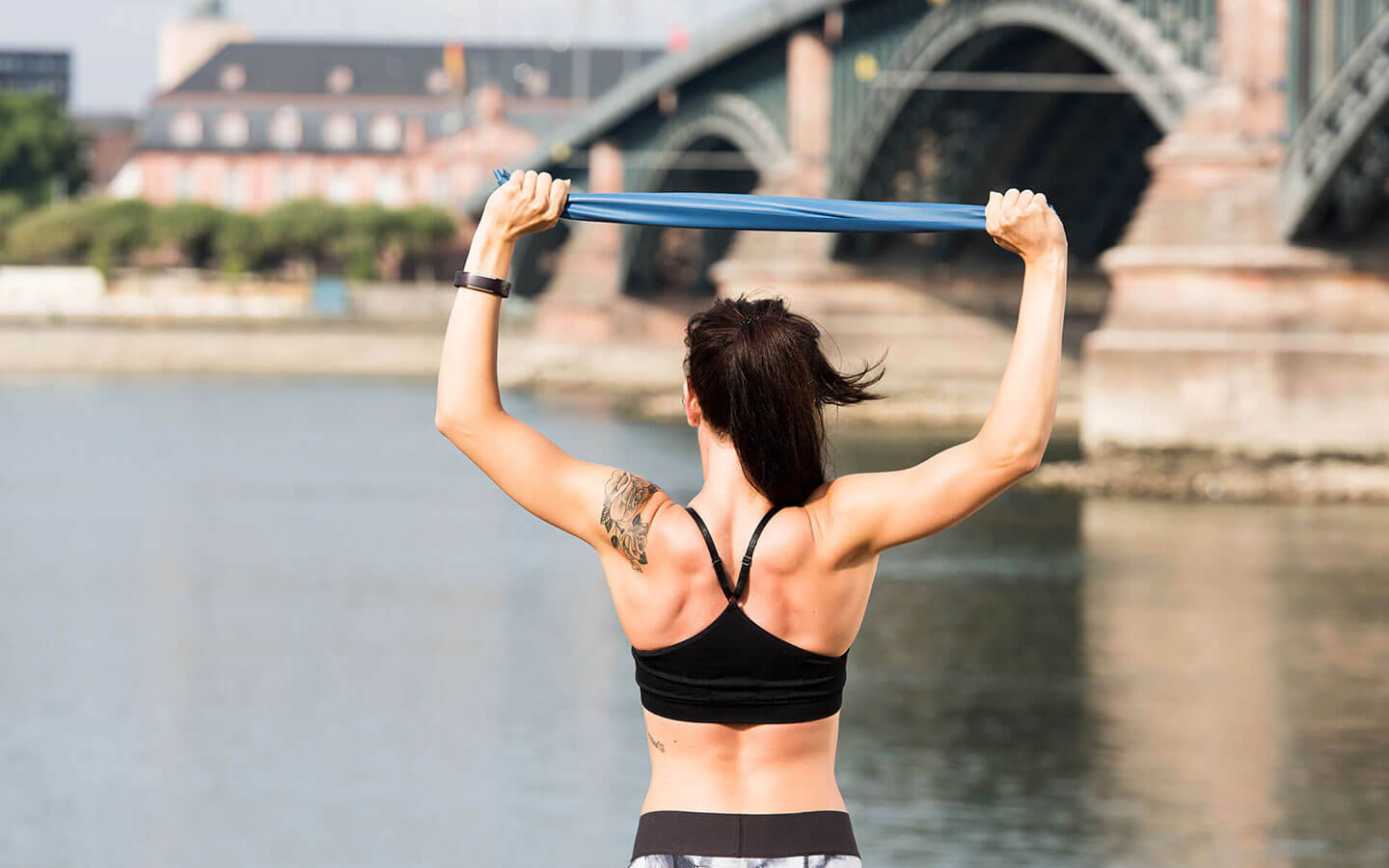 Frau steht mit dem Rücken zur Kamera an einem Flussufer und zieht über dem Kopf ein blaues Fitnessband mit beiden Händen auseinander.